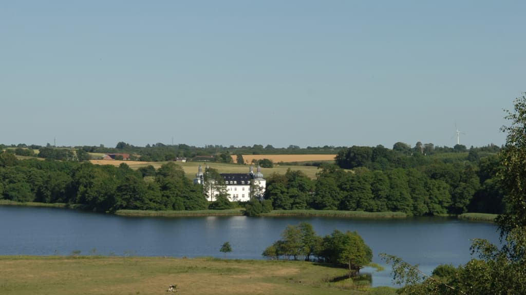 Engelsholm Schloss aus der Ferne mit Blick auf den See, die umliegenden Bäume und Felder.